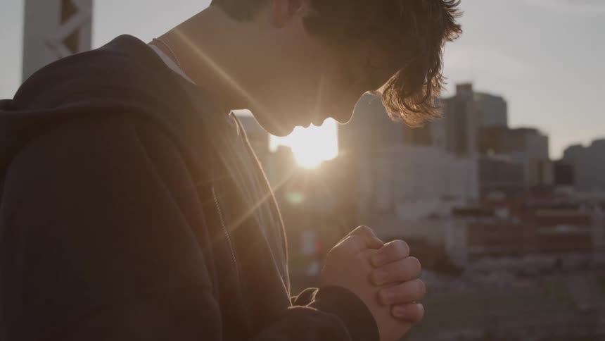 Young man bows his head and prays while the sun shines past the downtown buildings