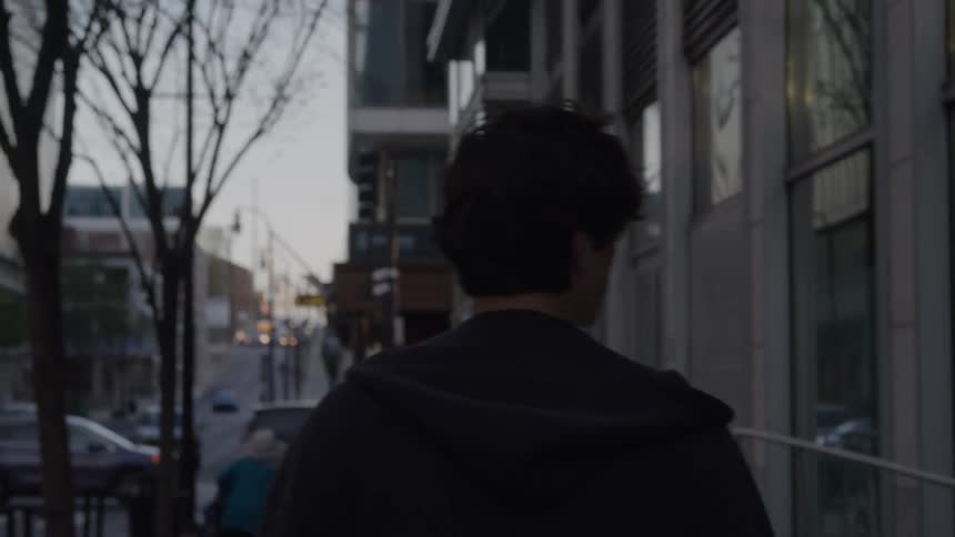 The video shows a persons back as they walk away down a city sidewalk lined with trees and tall buildingsthey are wearing a dark hoodie and the setting suggests an urban environment possibly in the late afternoon or early evening