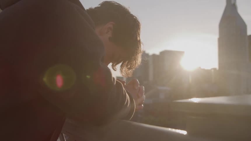 An young man wearing a dark hoodie leans on a railing with hands clasped looking down at a city skyline dominated by a tall building with a pointed roof with bright sun creating a flare behind him