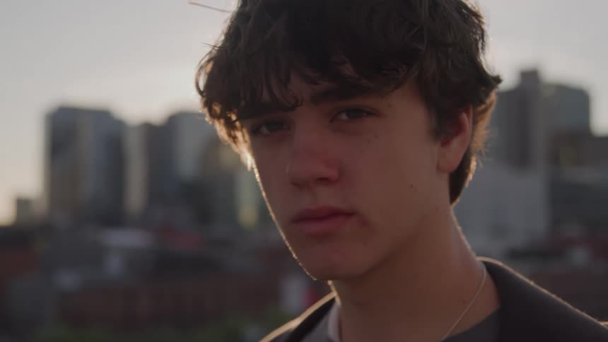 The video is a closeup shot of a young man with dark curly hair standing outdoors possibly on a rooftop in a citythe light suggests a sunset or golden hour as the sun casts a warm backlight that emphasizes his hair and silhouette against th
