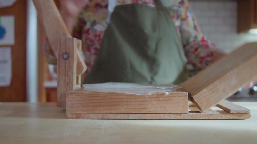 A close view shows a person in an apron and floral shirt pressing a dough ball with a sheet of plastic wrap in a wooden tortilla press then lifting out the flattened tortilla