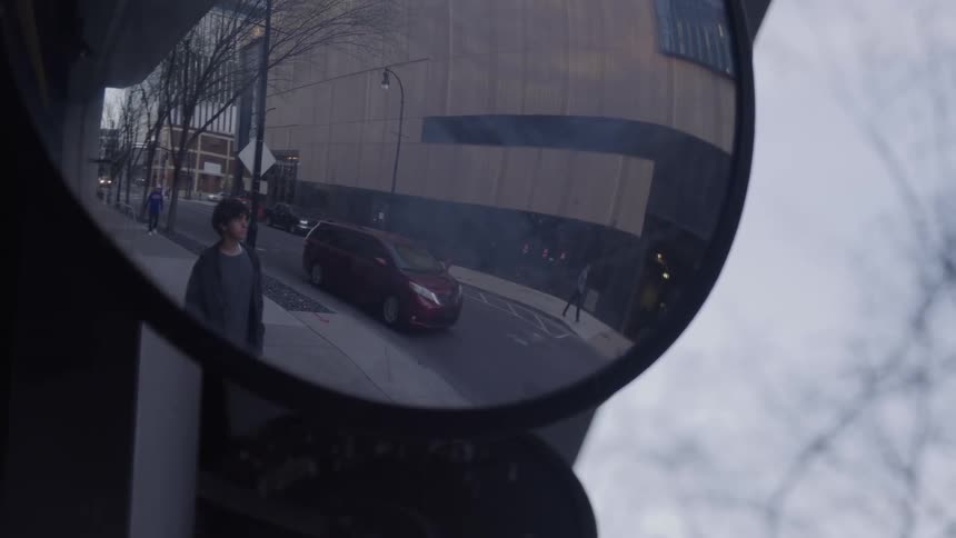 A wide angle convex safety mirror reflects a busy city street with a man walking on the sidewalk and cars driving past on an overcast day