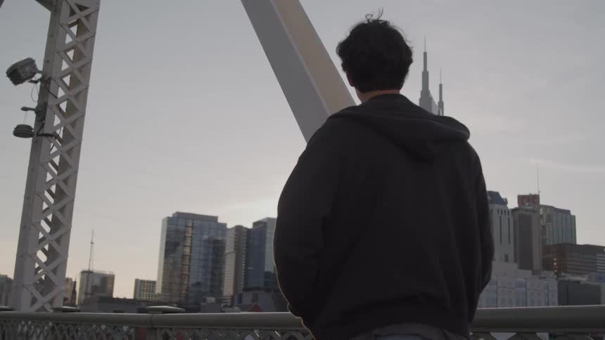 A young man wearing a jacket walks across a bridge at sunset with the city skyline visible in the distance