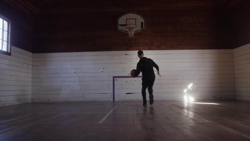 A man wearing dark clothing dribbles a basketball turns and shoots the ball toward a hoop all within a small wooden indoor court illuminated by light from the side windows
