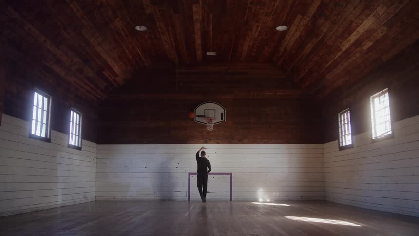 A full view of an old gymnasium shows a man wearing dark clothes shooting a basketball at a hoop mounted on a white wall under a high wooden ceiling with light streaming in from tall windows