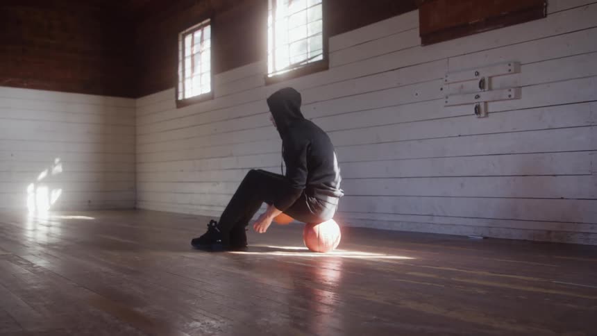 A person wearing a dark hoodie sits on an orange basketball on a wooden gym floor and bounces a second ball with bright sunlight streaming through two windows illuminating the floor and white walls