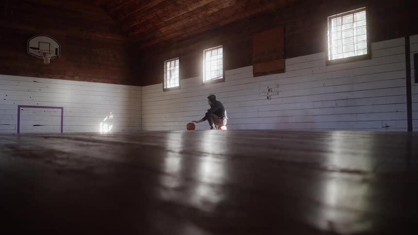 A person crouches low and repeatedly taps an orange ball on a polished wood floor silhouetted against a white wall with windows inside a long dimly lit indoor gymnasium with a woodplank ceiling