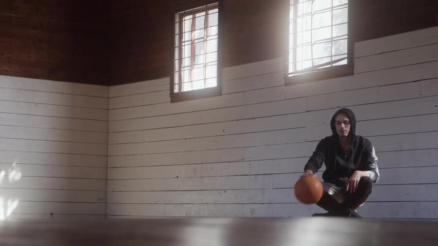 The video shows a young person sitting alone on the floor of a sunlit wooden building possibly a barn or shedwearing a dark hoodie they are holding a basketball on the ground while looking down in a quiet moment of contemplation