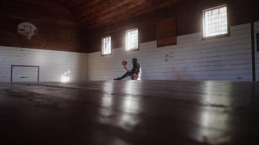 A man is sitting on an indoor basketball court and spinning a basketball on his finger