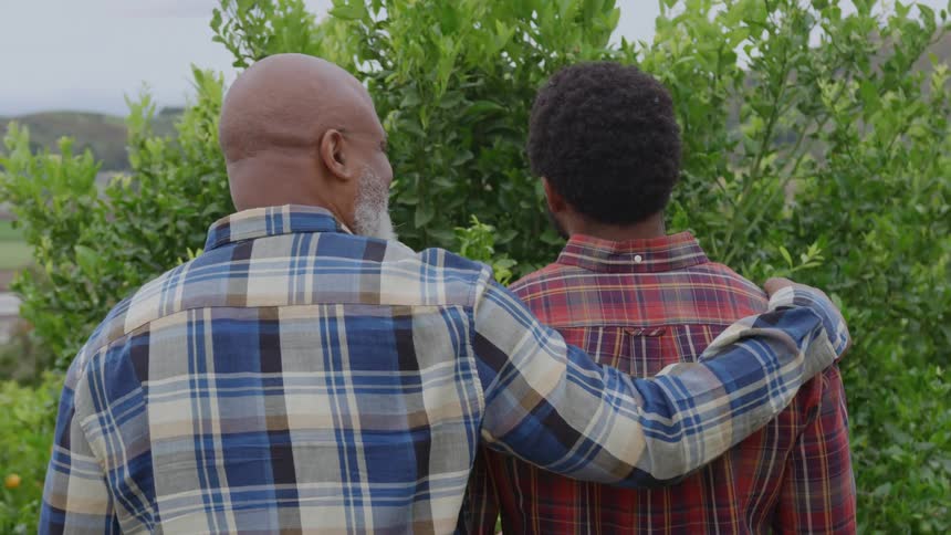 Two black men stand together watching a farmland covered with tall plants they smile as they talk and one rests his hand on the others shoulder