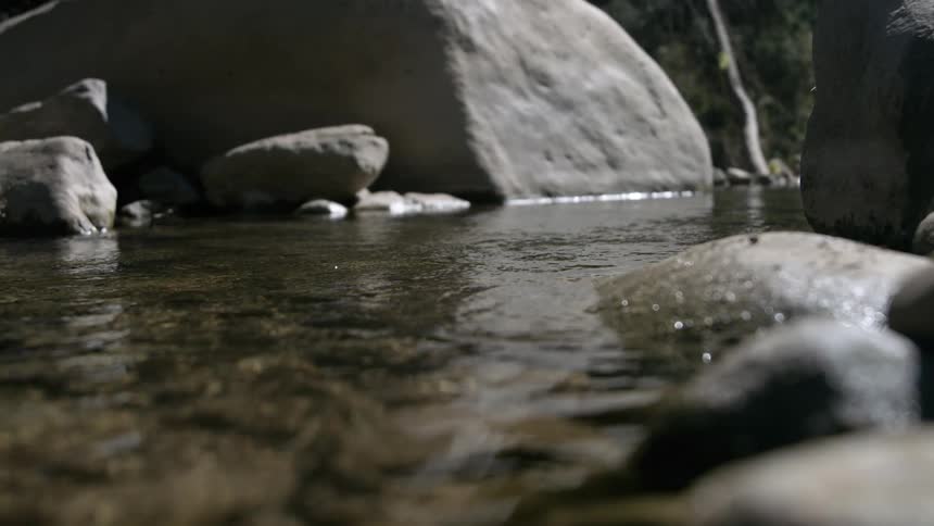Clear shallow river water flows gently over smooth river rocks creating small ripples and reflections against the dark backdrop of large smooth boulders