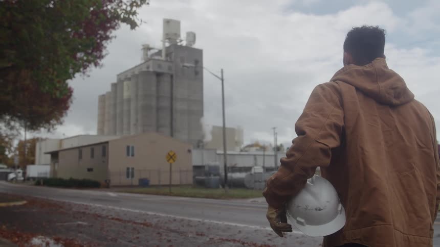 Worker holds his hardhat while standing near the road that leads past the nearby factory on a cloudy autumn day
