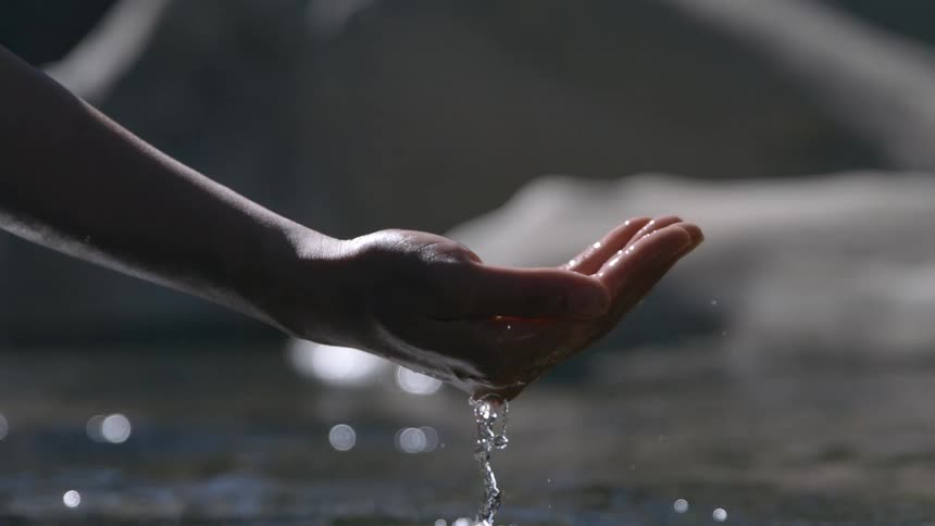 The video is a closeup shot of a hand cupping and collecting water as it drips down into the palmthe scene is backlit causing the hand and arm to be silhouetted against a bright hazy source of light