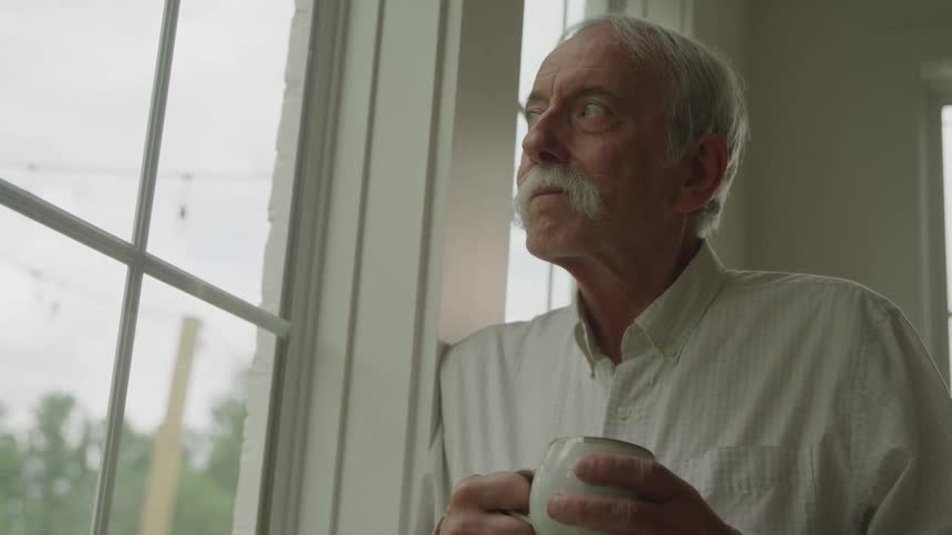 A closeup shot shows an older man with a mustache holding a white mug he stands indoors next to a large window looking outside at the cloudy sky and green trees