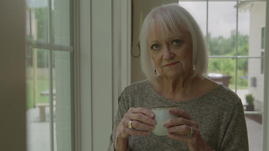 An elderly woman stands holding a coffee mug by a window indoors
