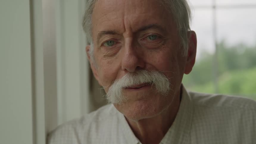 An elderly man with a mustache smiles while posing indoors