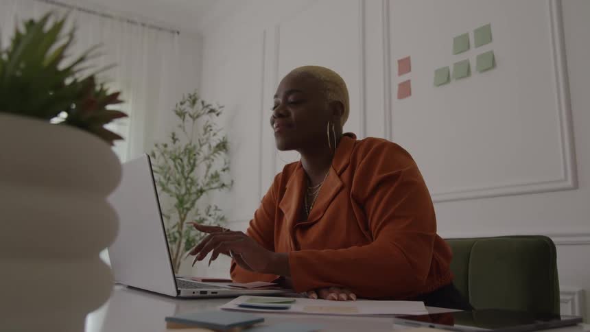 A woman with short blonde hair smiles while working at her desk transitioning from writing notes to typing on her laptop in a bright modern office