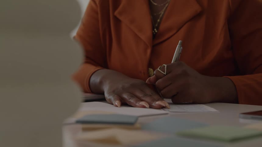 The video is a closeup shot of a womans hands wearing an orangebrown blazer and writing with a metallic pen on a stack of papers or notesher nails are manicured and painted a dark color as she focuses intently on signing or filling out a doc