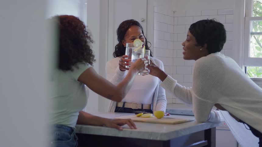 A trio of women in a bright kitchen clink glasses of lemon water around an island smiling