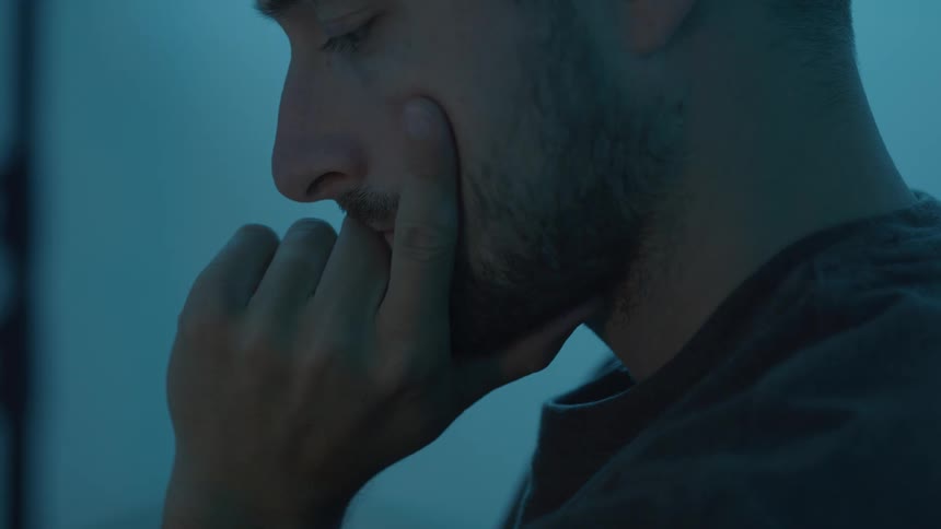 A man in cool blue light rests his face on his hand with a pensive expression