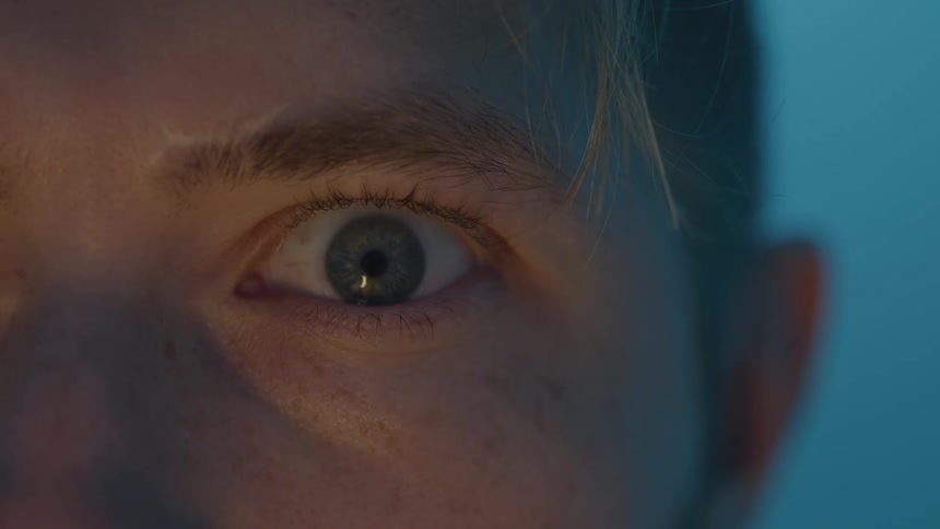 The video is an extreme closeup focusing on a closed eye in soft subdued lightinga subtle blue and orange light illuminates the skin suggesting the person might be looking at a screen or experiencing a specific ambiance