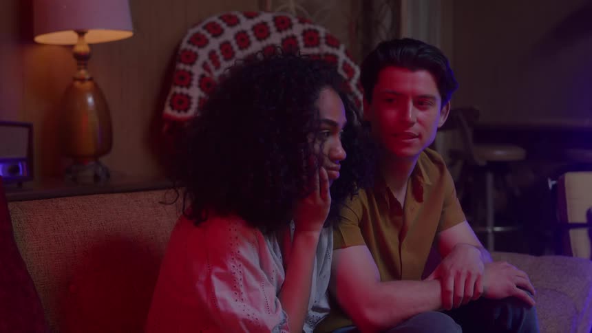 A man and woman sit closely together on a sofa in a dimly lit room the woman rests her chin on her hand while the man looks at her and then turns his head away with red and blue light illuminating the scene