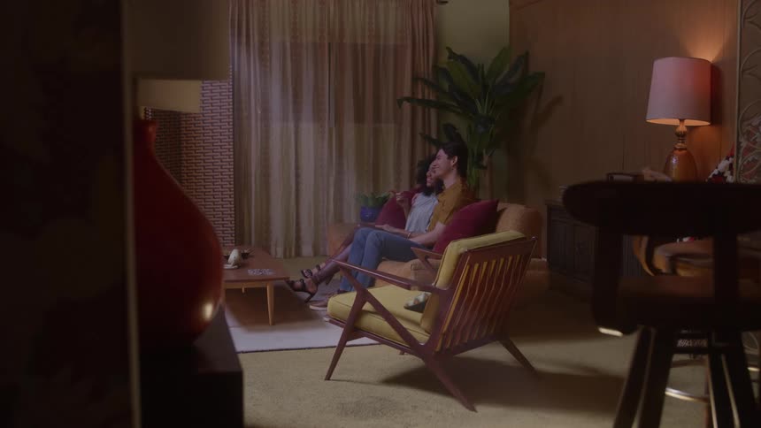 Two people sit on a sofa in a living room with dark walls and midcentury furniture leaning toward each other in conversation framed by an orange ceramic vessel and a wooden stool in the deep foreground