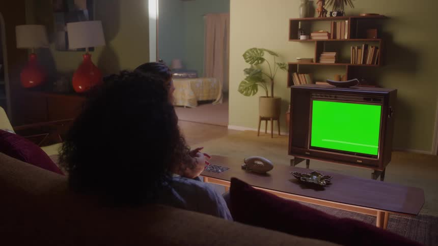A couple sits on a couch in a retro styled living room watching a vintage television with a green screen