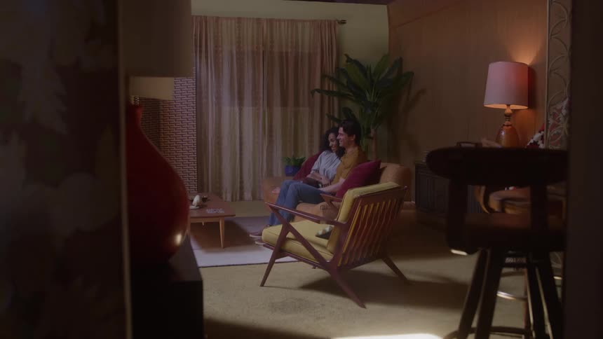A couple sits together on a midcentury chair in a cozy softly lit living room