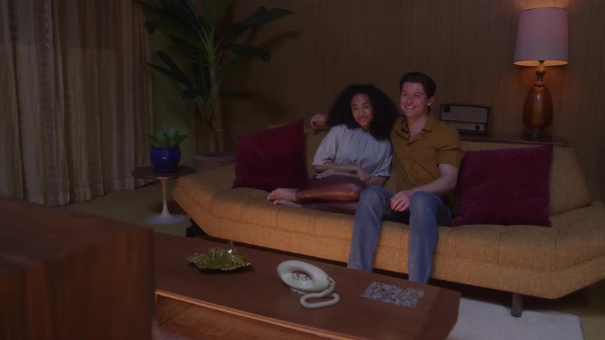 A young man sits on a couch watching a movie with a teenage black girl beside him resting his arm over her shoulder as they watch together