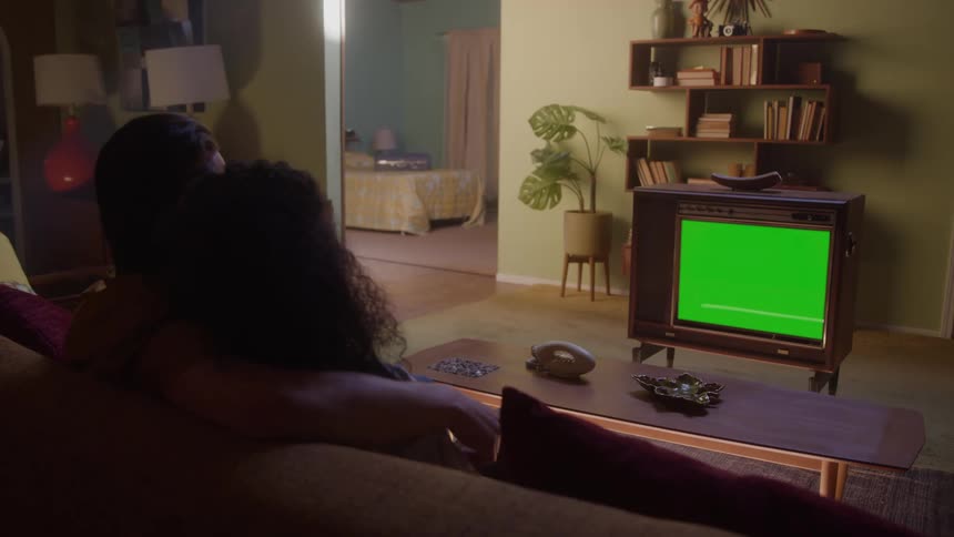 A couple cuddling on a sofa in a living room watching a television with a green screen