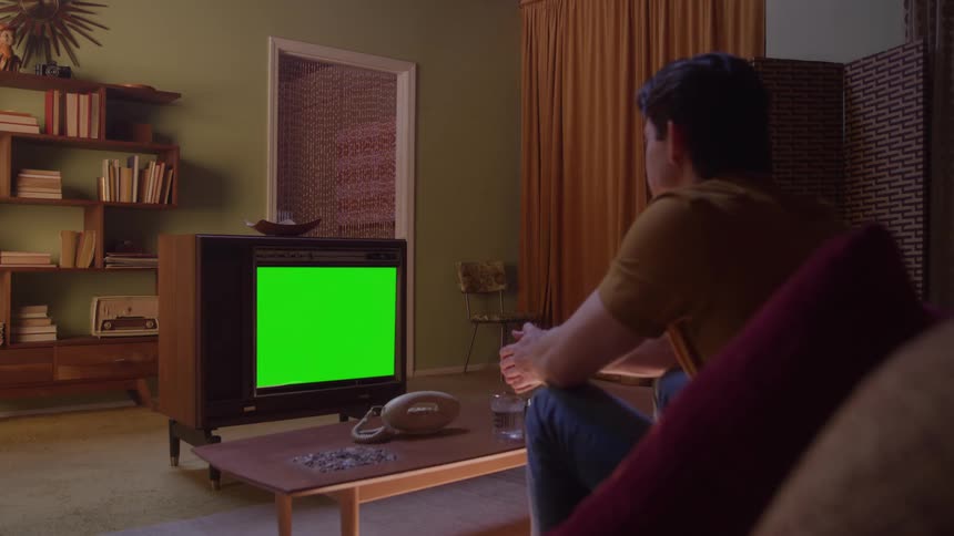 The video shows a man sitting on a retro sofa in a dimly lit vintagestyle living room relaxing and looking towards an old wooden tv with a green screen displayed the room features dark curtains olivegreen walls and midcentury furnitur