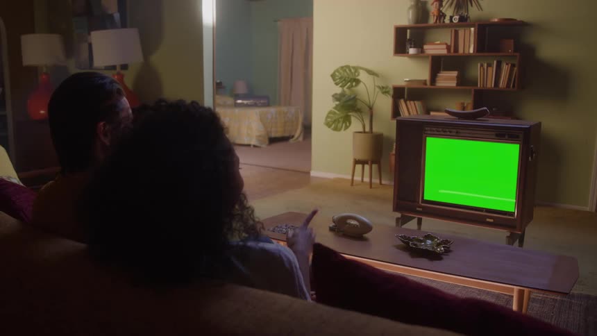 A man and a woman sit closely on a sofa their backs to the camera watching a television with a bright green screen the set placed in a living room with wooden furniture and shelves full of objects
