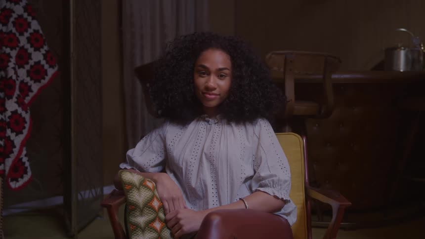 A young woman with long curly dark hair is seated in a vintagestyle yellow armchair in a dimly lit cozy room she is looking directly at the camera with a relaxed serene expression dressed in a lightcolored patterned blouse