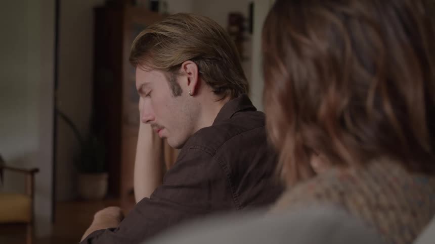 A medium closeup shot focuses on a man with blond hair looking down in distress as a womans hand rests reassuringly on his shoulder the two people are sitting indoors suggesting a private moment of comfort or concern within a warm domest