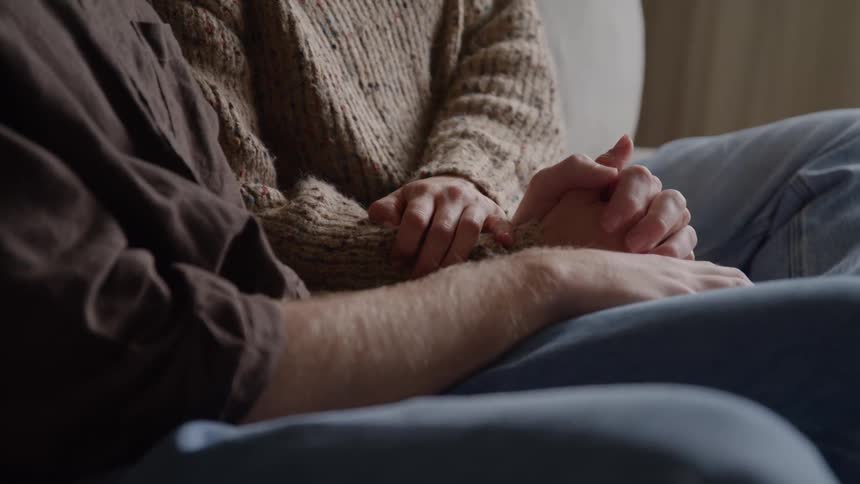 The video is a closeup shot focusing on the hands of a man and a woman clasped together as they sit next to each otherthe man wears a brown shirt and jeans while the woman is wearing a knit sweater suggesting a moment of comfort intimacy o