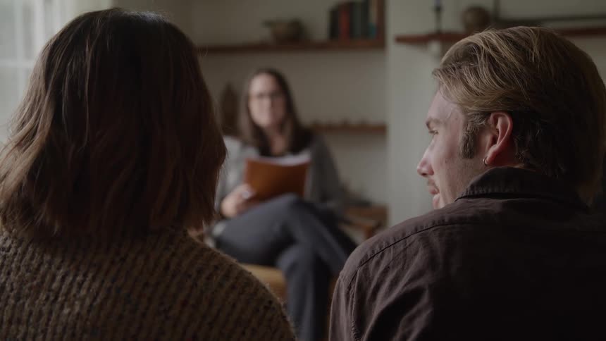 The backs of a man and a woman sitting in chairs face a therapist who sits with a clipboard the group engaged in discussion inside an office with a bookshelf in the background