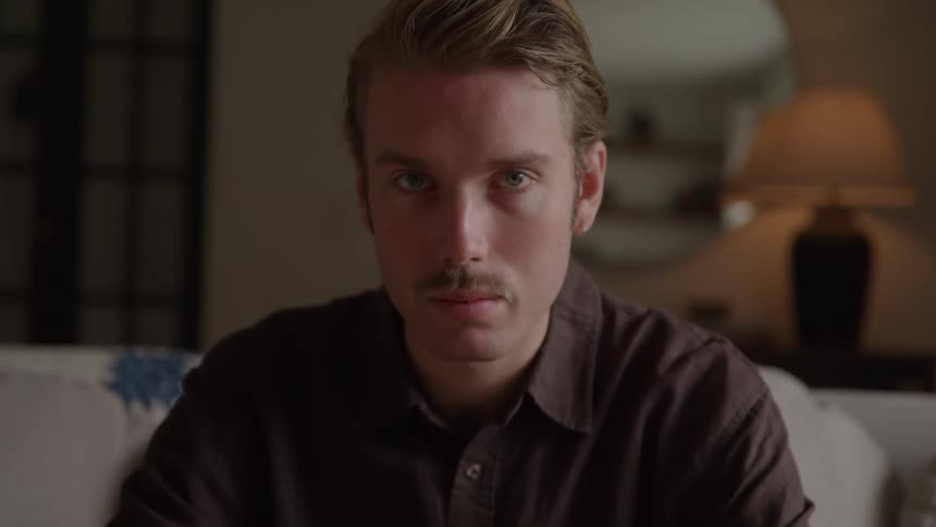 The video is an extreme closeup of a mans face illuminated by a soft warm light in a dimly lit roomhe has a mustache and is staring intently towards the camera with a serious or thoughtful expression
