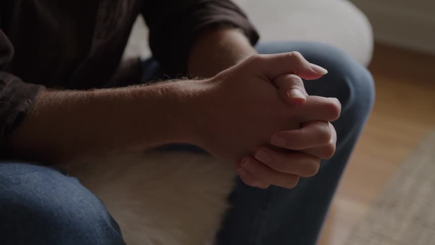A seated man in jeans clasps his hands together on his knee indoors appearing thoughtful