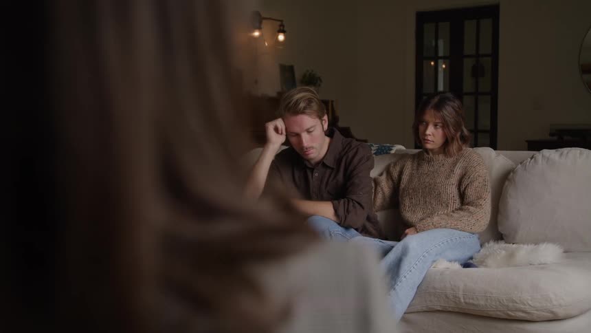 A couple is sitting on a white sofa in a living room and they have sad faces while someone in front is making hand gestures