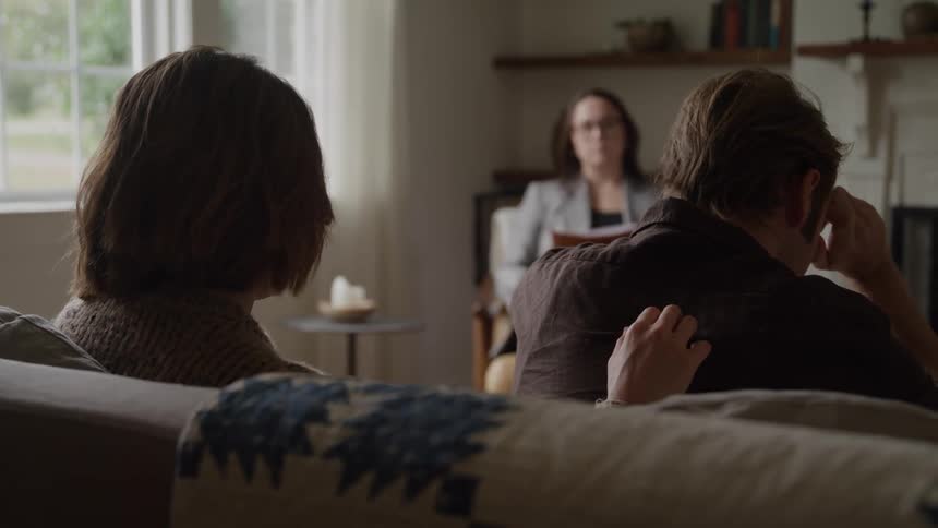 A man and a woman sitting on a sofa and the woman is stroking his back while the man is in sad tones holding his head with his hand