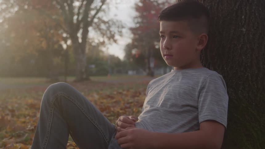 Boy sighs while sitting on the leaf covered ground and leaning against a tree on an autumn day
