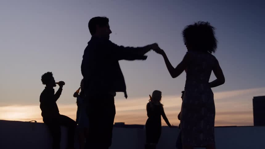 A group of people are silhouetted against a colorful sunset sky on a rooftop as a man and woman stand close and dance together at a casual party