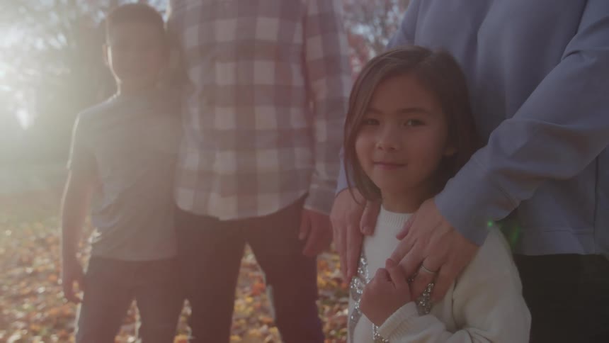 This is a warm brightly lit shot of a family standing outside likely in the fall with dappled sunlight creating lens flare in the background the focus is on a young girl smiling at the camera while an adults hand rests protectively on her sho