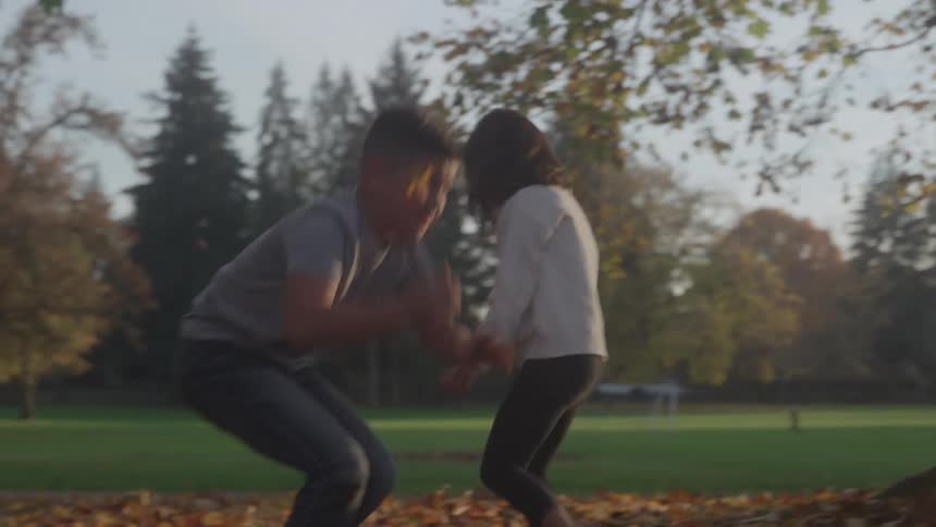 Two children run and throw brown and yellow fallen leaves in a park setting surrounded by trees and grass under the light of a setting sun and an arm of a third person is visible