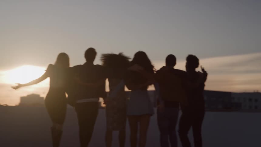 The video is a sunsetlit silhouette of a group of six friends standing shouldertoshoulder on a rooftopthe group is celebrating with arms around one another and one person raises their hand in a peace sign gesture