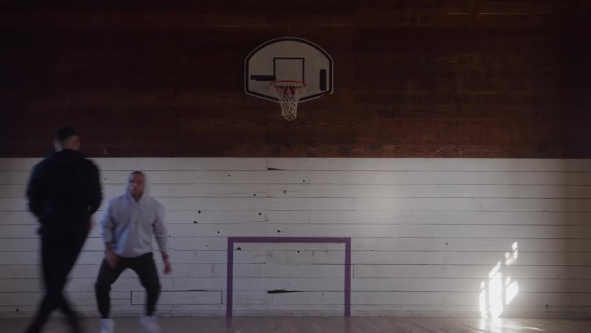 The video is an interior shot of a wooden gymnasium or studio where a man is dribbling a basketballhe is dressed in dark clothing and is standing in the middle of the frame facing a basketball hoop mounted on a wooden wall