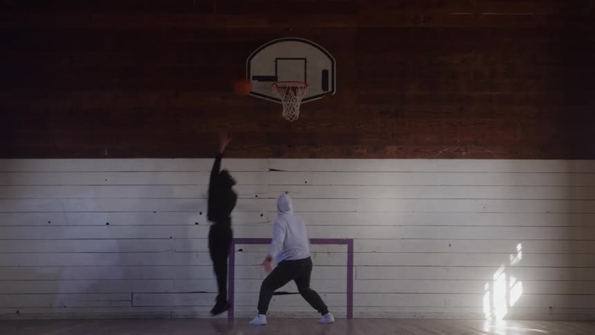 Two men play basketball on a wooden indoor court beneath a hoop