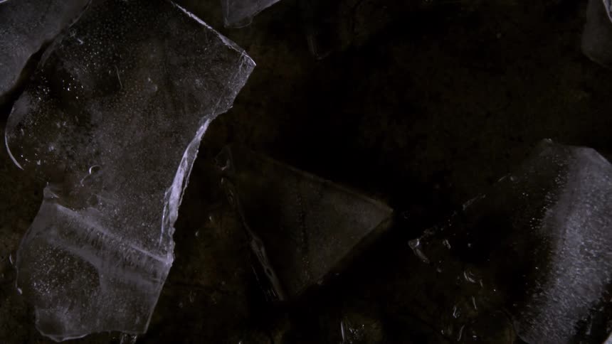 A closeup view shows several broken pieces of clear ice resting on a dark surface as the ice slowly melts the water drips and runs off the surface