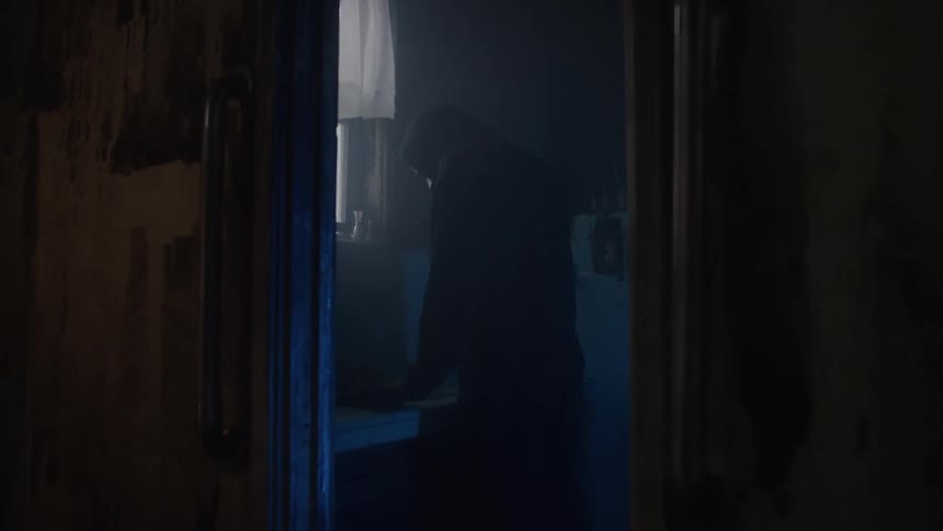 A dark hooded silhouette of a person is framed by a doorway with wooden sides and metal handles the man stands in a bright inner room